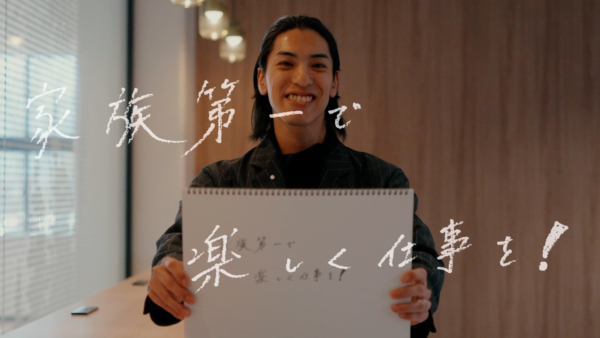 調達のKさんが「家族第一で楽しく仕事を！」と書いた手書きのフリップを持ち笑顔を見せる様子